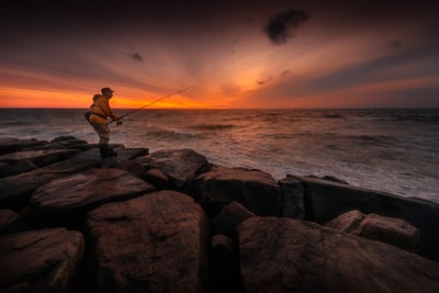 日落时站在岩石上在水面钓鱼的人
